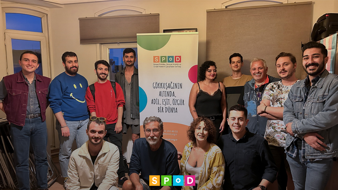 SPoD ofisinde düzenlenen genel kuruldan kalabalık bir grup fotoğrafı. Kişilerin ortasında "gökkuşağının altında adil, eşit bir dünya" sloganı yazan bir kırlangıç var. En altta SPoD logosu mevcut.